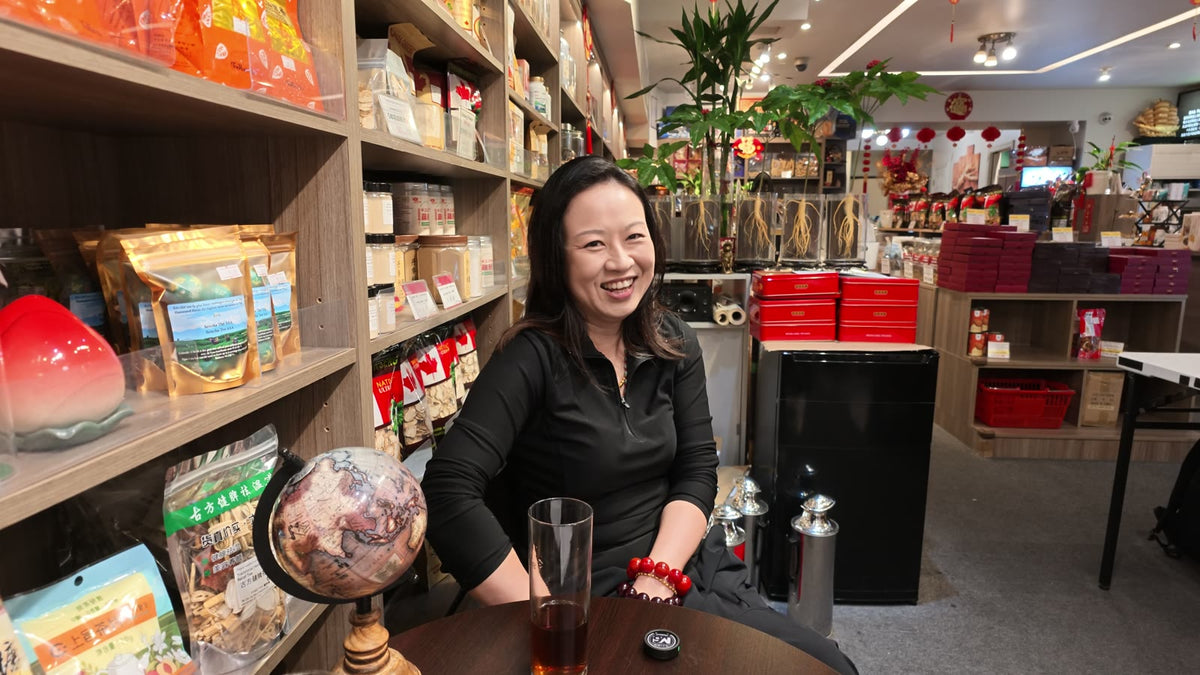 Carmen smiling during an interview at a small table inside Fosetter, with shelves of ginseng and dried goods around her.