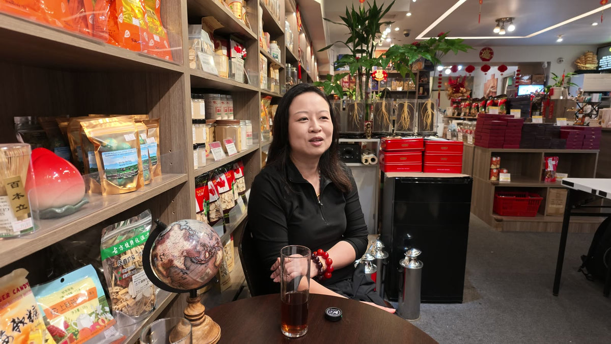 Carmen talking at a table inside Fosetter, with ginseng packages and a globe on the shelves beside her.