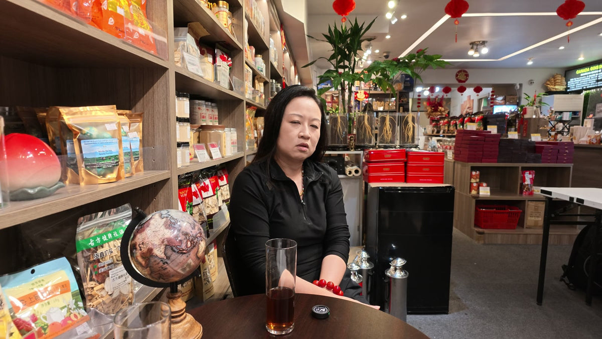 Carmen seated at a table inside Fosetter, with shelves of ginseng and dried goods behind her.